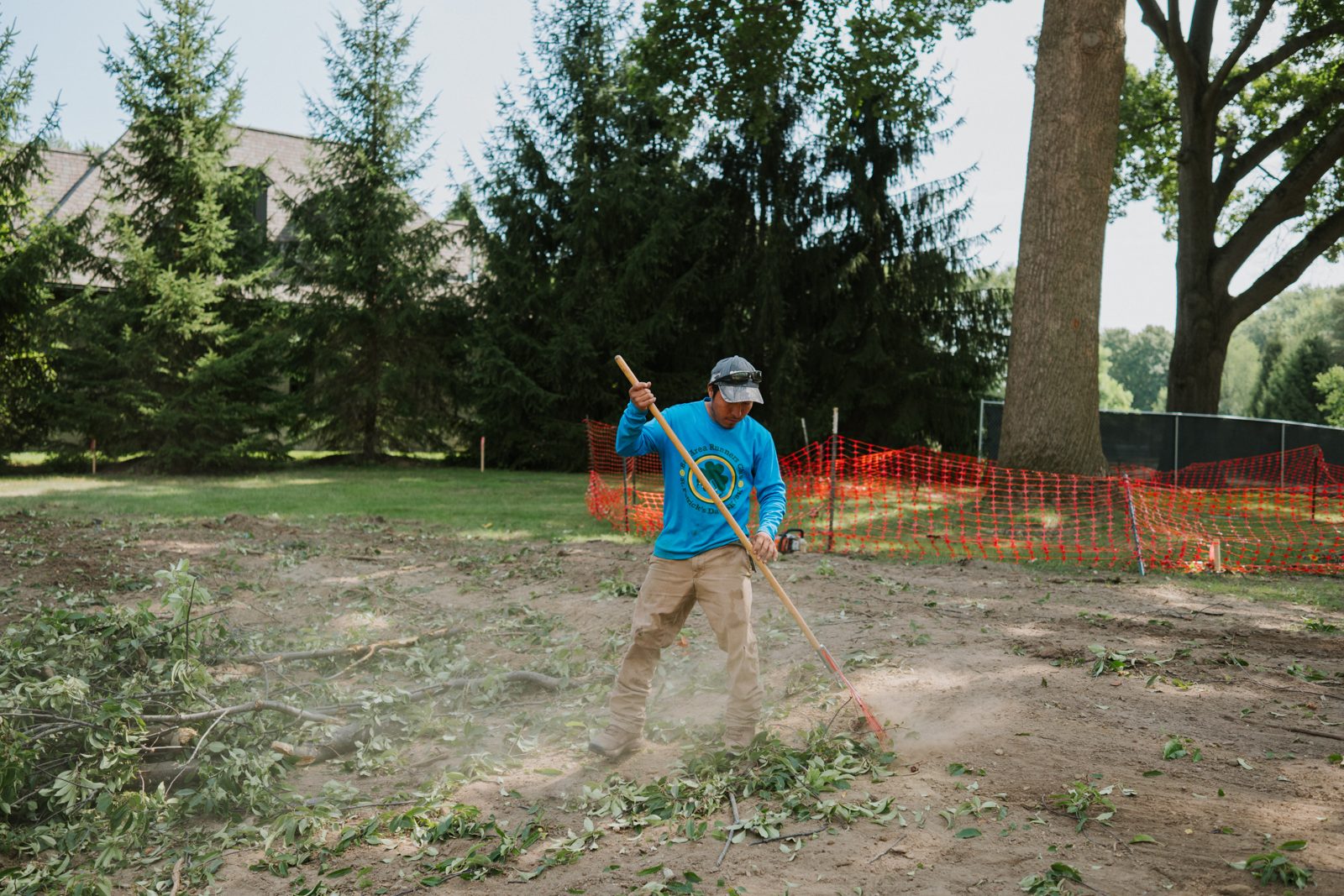 Crew member raking debris on a cleared site with tall spruce trees behind him